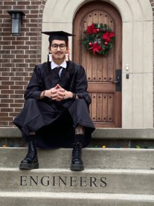 man in graduation gap and gown sitting in front of door